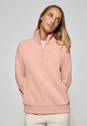 Lichtroze ritsvest met hoge kraag, geribbelde onderrand en subtiele logodetails. Gemaakt van zachte stof met een gladde textuur.
