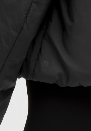 Detaljni prikaz rukava i bočne strane crne puf jakne s diskretnim okruglim Lululemon logotipom na tkanini.