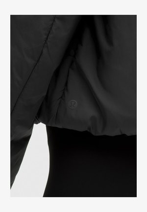 Detaljni prikaz rukava i bočne strane crne puf jakne s diskretnim okruglim Lululemon logotipom na tkanini.