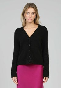 Cardigan noir en maille avec un col en V, fermeture par boutons à l'avant, poignets et ourlet côtelés. Porté sur une jupe rose. Texture douce.