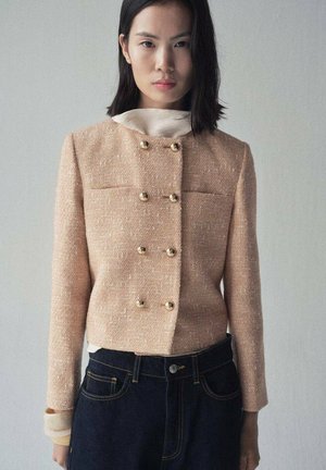 Peach tweed blazer with double-breasted front and gold buttons, featuring a cropped style and long sleeves, paired with dark denim jeans.