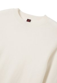Licht beige sweater met een geribbelde ronde hals en textuurstof. Bevat een klein zwart en rood label bij de hals. Geen zichtbare patronen of accenten.