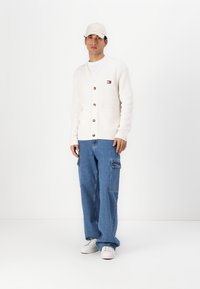 Kremfarget ribbestrikket cardigan med treknapper og frontlomme, brukt over en hvit T-skjorte; avslappede blå cargo-jeans og hvite joggesko.