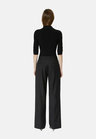 Woman standing facing away wearing a black long-sleeve top and high-waisted wide-leg black pants against a plain light background.