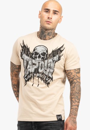 Helles beiges T-Shirt mit einem großen schwarzen Totenschädel-Graphic und dem Text "TAPOUT" sowie Flügeln. Hergestellt aus weicher Baumwolle mit kurzen Ärmeln.
