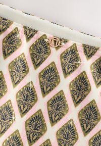 Tessuto con motivi di foglie verdi e nere a forma di rombi su sfondo rosa pallido e bianco, con un piccolo ciondolo rotondo dorato attaccato.