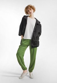 Osoba z kręconymi włosami ubrana w czarną kurtkę, białą koszulę, zielone spodnie i białe trampki, stojąca z rękami w kieszeniach na białym tle.