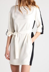 Robe blanche avec des panneaux latéraux noirs, manches trois-quarts et taille nouée. Texture lisse, design minimaliste et poches latérales.