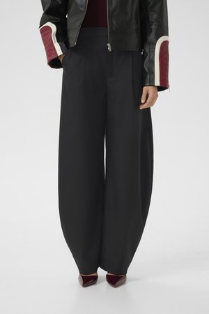 Woman wearing black wide-leg pants, black leather jacket with red and white sleeve patches, and dark red pointed shoes.