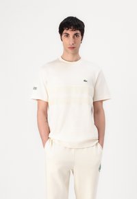 Cremefarvet bomulds T-shirt med ribbet rund hals, korte ærmer og et horisontalt stribet design i en lysere tone, med et grønt logo.