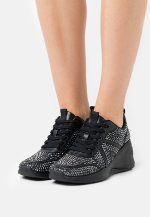 Baskets noires à semelles épaisses, ornées de clous argentés, portées pieds nus sur un fond blanc.