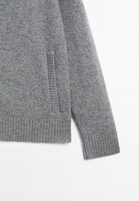 Maglione grigio con una texture morbida, dotato di polsini e orlo a costine, e una tasca laterale integrata nel tessuto a maglia.