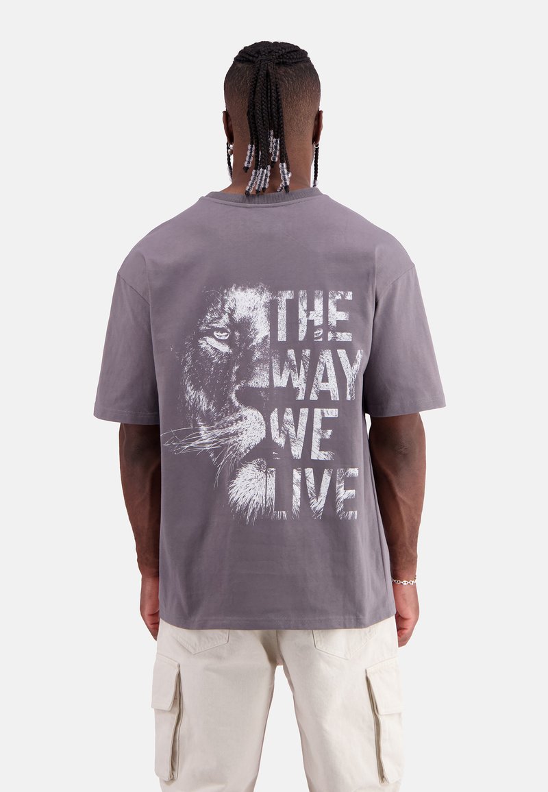 Graues Baumwoll-T-Shirt mit einem auffälligen weißen Aufdruck eines Löwenkopfes und dem Text "DER WEG, WIE WIR LEBEN" auf dem Rücken. Kurze Ärmel, lockerer Schnitt.