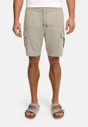 Mann trägt beige Cargo-Shorts mit Kordelzugbund und graue Sandalen mit doppeltem Riemen, steht vor weißem Hintergrund.