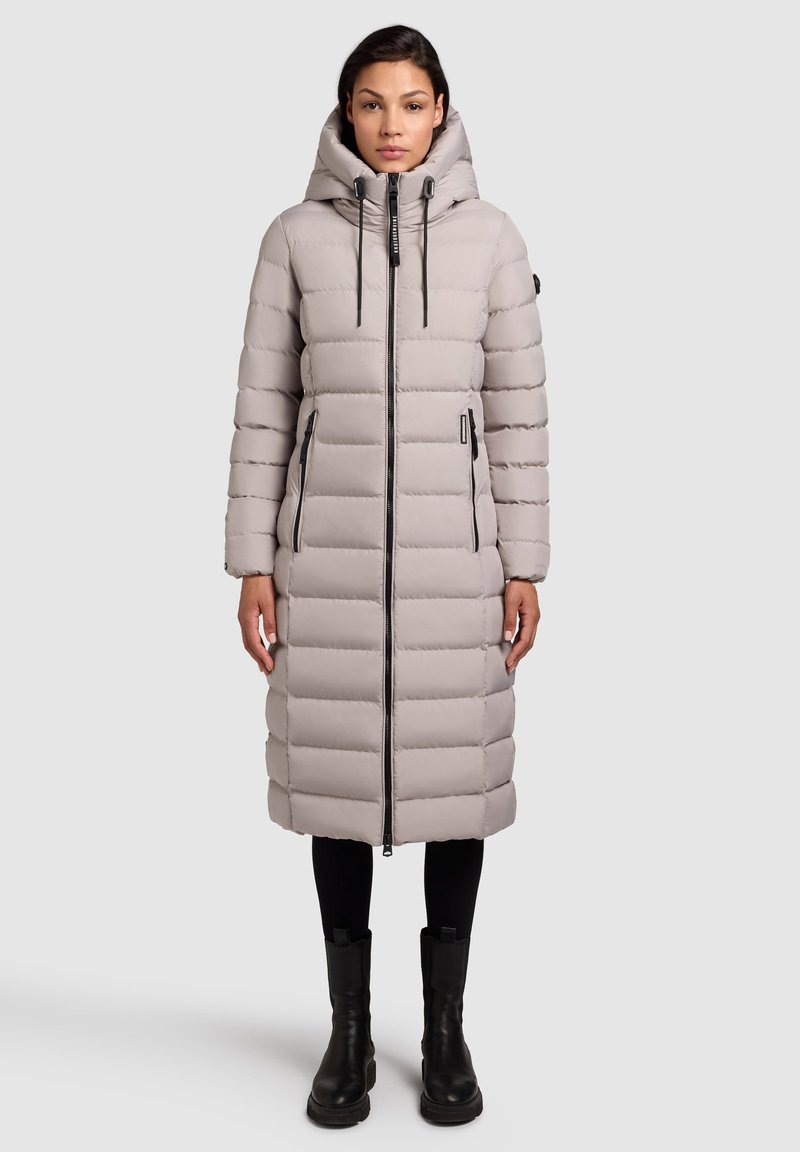 Lys grå lang dunjakke med hætte, der har vandrette quiltede mønstre, en frontlynlås og to sidelommer. Sorte accenter og støvler.
