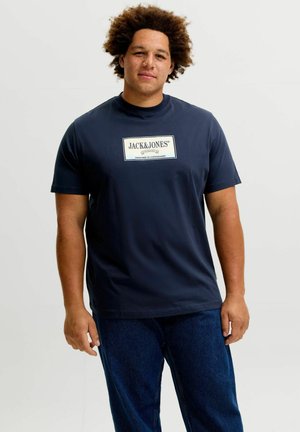 Junger Mann mit lockigem Haar, der ein marineblaues Jack & Jones T-Shirt und dunkelblaue Jeans trägt, vor einem schlichten hellen Hintergrund stehend.