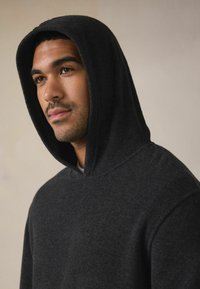 Mørkegrå hættetrøje lavet af tekstureret stof med en stor hætte og afslappet pasform, med synlige sømme ved halsudskæringen og ærmerne.