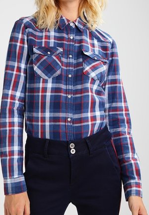 Person trägt ein blaues, rotes und weißes kariertes Hemd mit Knöpfen, zwei Brusttaschen und dunkelblaue Hose mit doppelten vorderen Knöpfen.