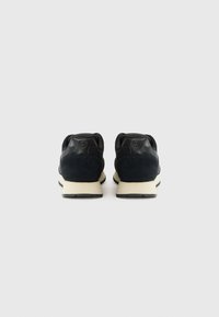 Schwarze Wildleder-Sneaker mit Lederakzenten und einer beigen Zwischensohle. Abgerundetes Toe-Design und Gummisohle, die von der Rückansicht sichtbar ist.