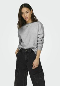 Szürke pulóver hosszú ujjakkal, laza fitten, laza fekete cargo nadrágokkal párosítva, amelyek oldalzsebekkel és gombos zárással rendelkeznek.