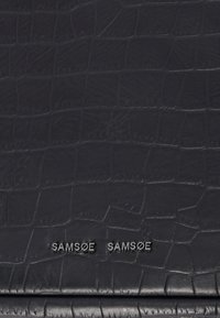 Cuir noir embossé façon crocodile avec une texture lisse. Présente une marque embossée "SAMØE SAMØE" en une police serif élégante.