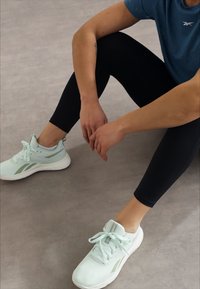 Világoskék sportcipő fehér talppal, zöld díszítésekkel és fűzőkkel. Fekete leggings-szel és kék felsővel viselve.