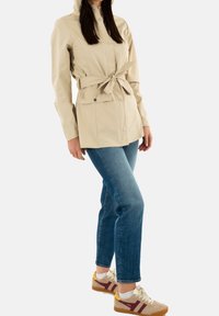 Veste imperméable beige avec ceinture ajustable à la taille, boutons-pression et capuche, associée à un jean bleu et des baskets beiges avec des accents bordeaux.