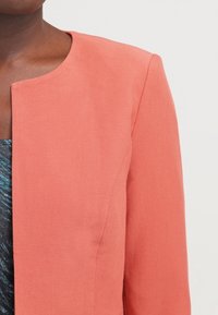 Blazer couleur pêche avec un col rond, une coupe ajustée et un tissu texturé ; présente des lignes épurées et un design minimaliste.