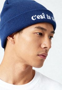 Bonnet tricoté bleu avec une texture côtelée, arborant le texte brodé en blanc "c'est la vie" sur le revers. Porté par-dessus une chemise blanche.
