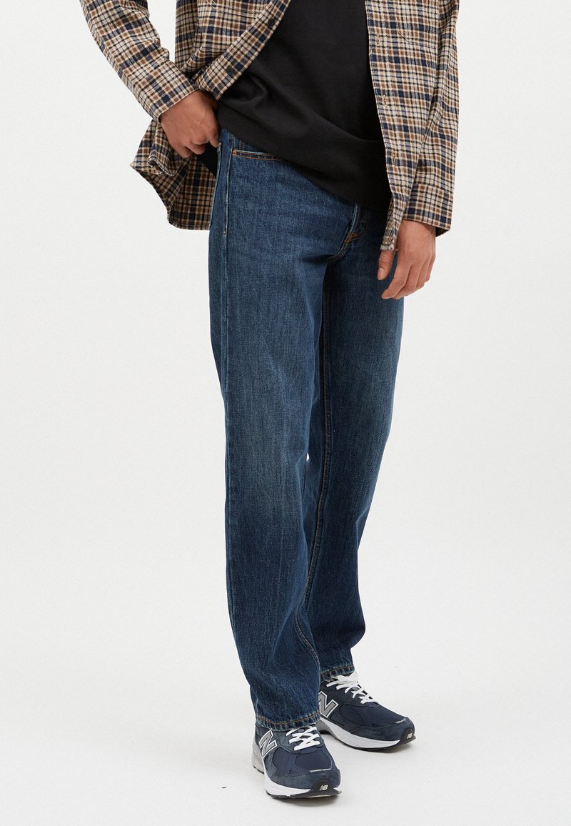 Vaqueros de mezclilla de un azul oscuro con corte recto, que cuentan con cinco bolsillos y un ligero desgaste. Combinado con una camiseta negra y una camisa de cuadros.