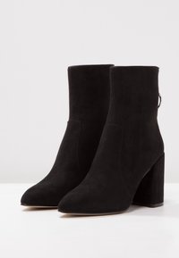 Bottines en daim noir avec un bout pointu, un haut talon bloc et des coutures minimales. Comprend une languette à l'arrière pour un enfilage facile.