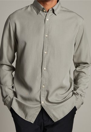 Uomo che indossa una camicia a maniche lunghe grigio chiaro con colletto e bottoni, mani nelle tasche, abbinata a pantaloni scuri su sfondo neutro.