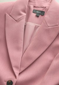 Blazer rose en tissu texturé, avec revers crantés et un bouton noir unique. L'intérieur est d'un rose plus clair avec une finition lisse. Étiquette de marque visible.