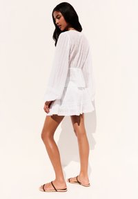 Femme aux longs cheveux noirs, vêtue d'une robe blanche à manches longues texturée et de sandales à brides noires, se tenant devant un fond clair et uni.