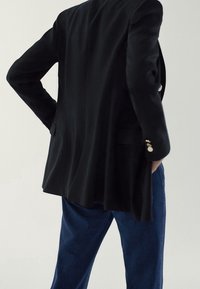 Blazer noir ajusté avec une texture lisse, doté de boutons dorés et de deux poches latérales, associé à un pantalon en denim bleu foncé.