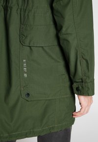 Grön tygjacka med vertikala fickdetaljer, kontrasterande sömmar och knäppning vid ärmarna. Slät textur och en avslappnad, rak silhuett.