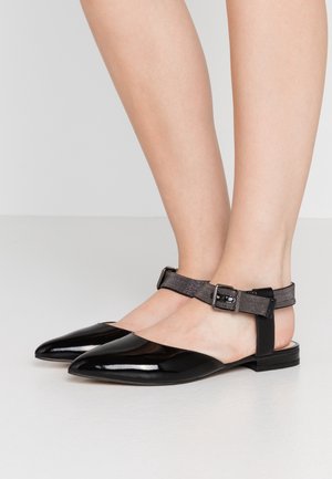 Füße, die glänzende schwarze Ballerinas mit spitzen Zehen und dunkelgrauen Knöchelriemen sowie einer silbernen Schnalle auf einem hellen Boden tragen.