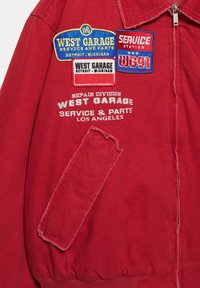Červená bunda s nášivkami s nápisy "West Garage Service and Parts Detroit Michigan" a "Service Station WG-91" a se šikmým kapsou vpředu.