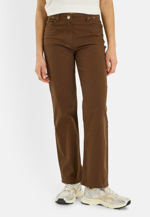 Femme portant un pantalon marron taille haute à jambes droites et des baskets blanches, debout devant un fond uni.