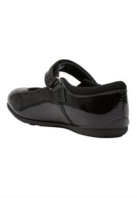 Scarpa in vernice nera con punta tonda, cinturino alla caviglia e suola in gomma bassa. Presenta decorazioni brogue sulla parte superiore.