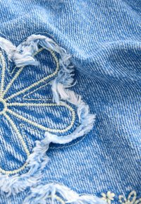 Tissu en denim bleu avec des bords effilochés et des broderies florales en crème. La texture est douce, avec des coutures détaillées visibles sur le fond bleu.