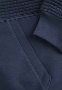 Navyblauer Hoodie mit strukturiertem Oberteil, glattem Stoff, gerippten Ärmeln und Seitentasche mit sichtbaren Steppnähten.