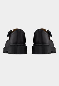 Ζευγάρι μαύρα δερμάτινα υποδήματα χαμηλής κοπής με χοντρές αυλακωτές σόλες και λουριά με αγκράφες στο πλάι, από πίσω σε λευκό φόντο.