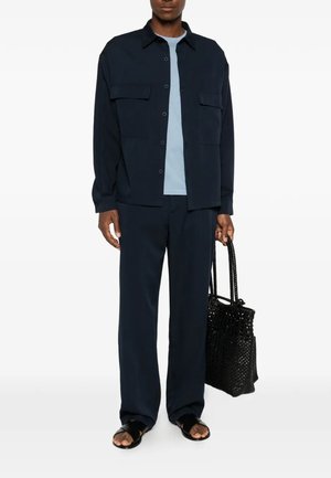 Adulto che indossa giacca blu navy con bottoni e pantaloni con camicia azzurra, tiene una borsa intrecciata nera e indossa sandali neri.