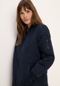 Giacca a vento navy con un design a trapunta a diamante, zip frontale, polsini a coste e tasca laterale con dettaglio a zip. Leggera e dalla texture piacevole.