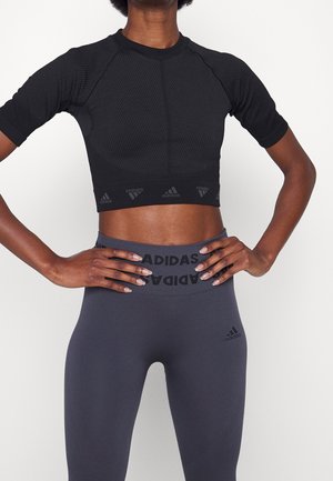 Sportowiec w czarnym krótkim crop topie i szarych legginsach z wysokim stanem z logo Adidas, ręce spoczywające na biodrach na białym tle.