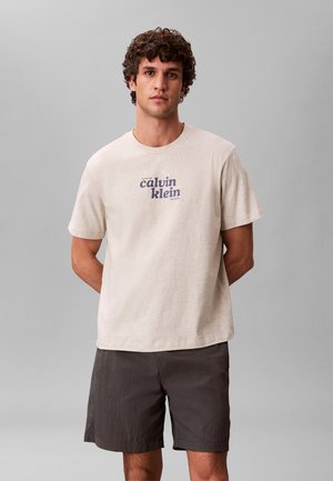 Mężczyzna o kręconych włosach, ubrany w beżowy t-shirt Calvin Klein i ciemnoszare szorty, stoi z rękami za plecami na jednolitym tle.