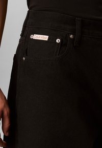 Nahaufnahme einer schwarzen Denim-Jeans-Tasche mit Calvin Klein Etikett und Metallnieten, getragen von einer Person mit schwarzem Oberteil.