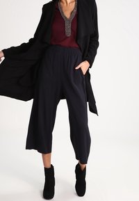 Manteau long noir sur un haut bordeaux, associé à des culottes larges bleu marine et des bottines noires ; présente un col orné et des poches.