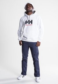Sudadera blanca con capucha y logo de "Helly Hansen" en rojo y negro, pantalones azul marino y zapatillas blancas. El modelo lleva un sombrero de cubo blanco.